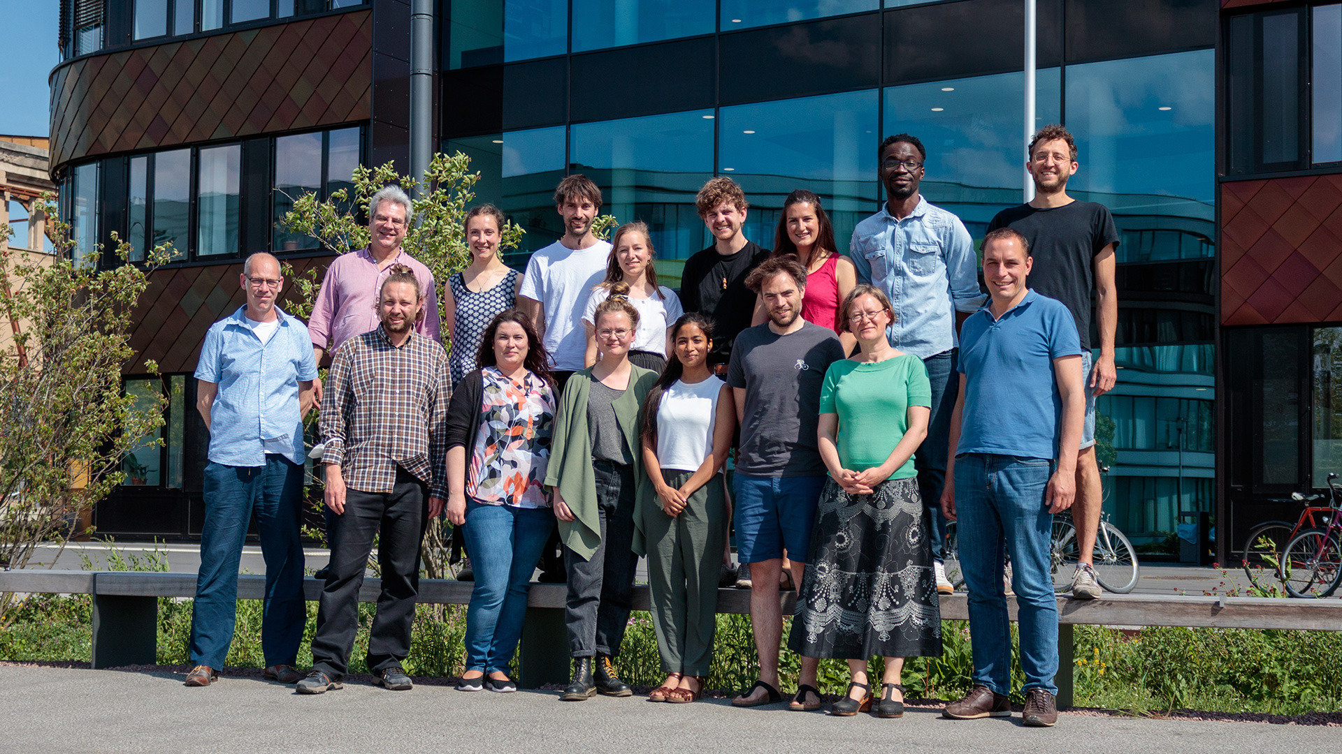 idiv-A group of fifteen people stand outdoors in front of a modern building, smiling at the camera. They are dressed casually, and the day appears sunny and clear.